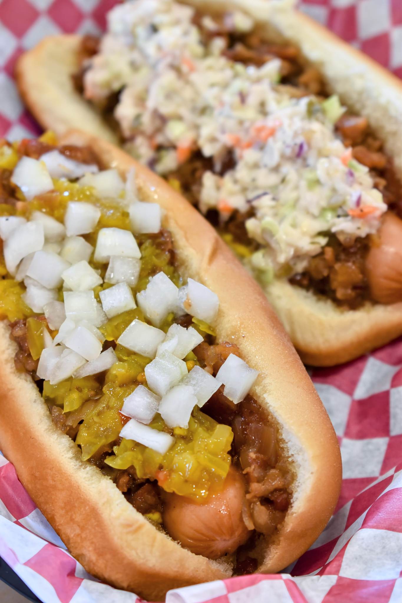 Two loaded deluxe hot dogs in a basket with chili, chow chow, slaw, and diced onion